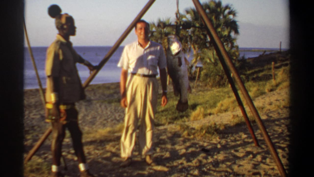 1969 Sudan home-movie frame showing a white-clothed man posing beside a large fish on a tripod while another adult stands nearby.