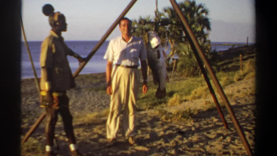 1969 Sudan home-movie frame showing a white-clothed man posing beside a large fish on a tripod while another adult stands nearby.