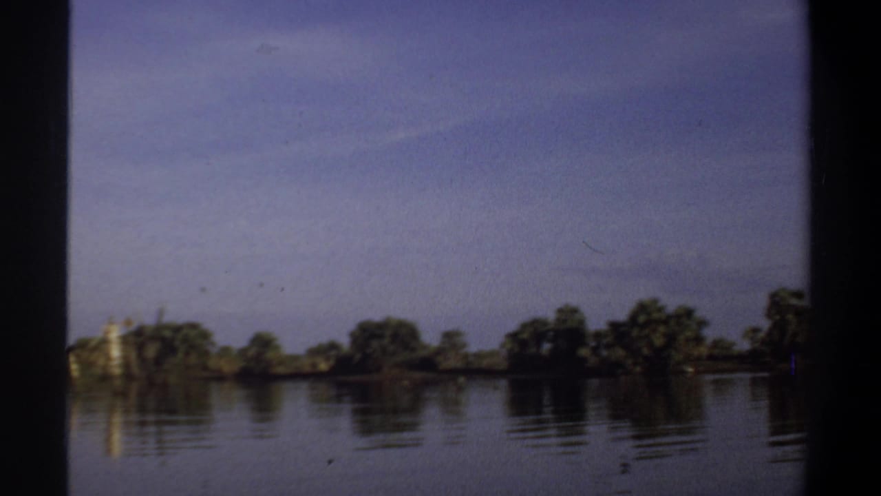 1969 Khartoum home-movie frame showing calm water, distant shoreline trees, and soft reflections under a blue sky.