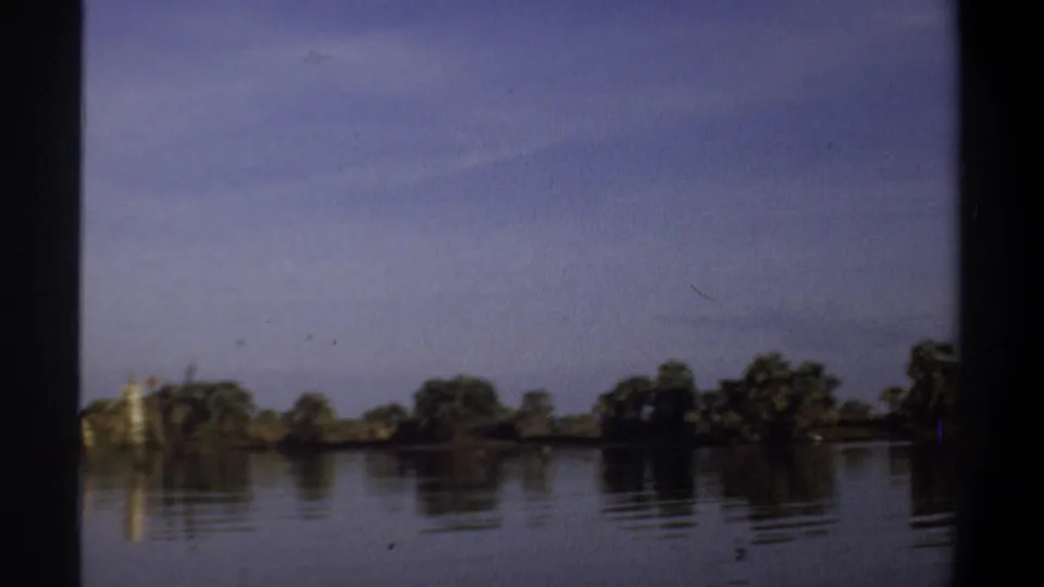 1969 Khartoum home-movie frame showing calm water, distant shoreline trees, and soft reflections under a blue sky.