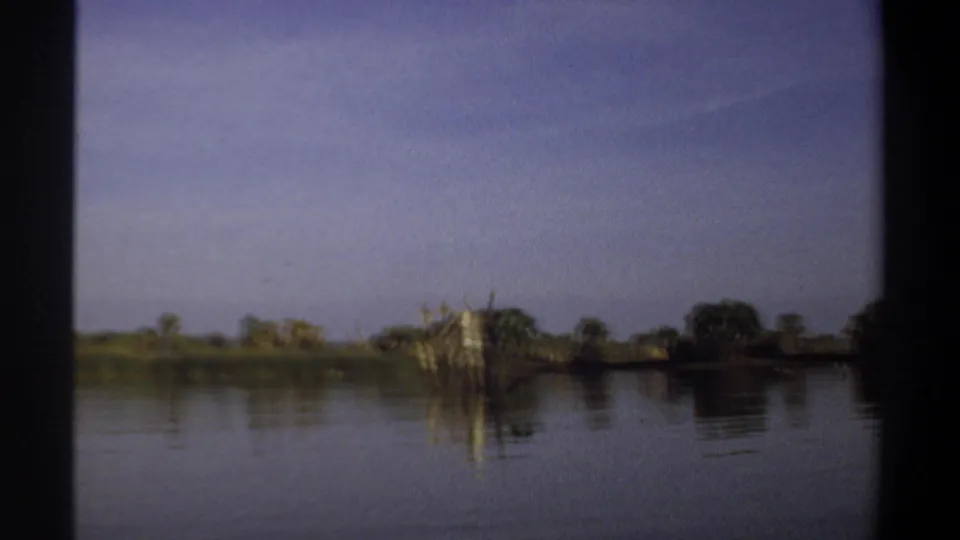 1969 Khartoum river view with still water, low shoreline vegetation, and weathered wooden posts rising near the right side.