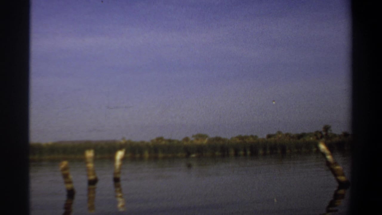 1969 Khartoum-area home-movie frame showing calm river water, wooden posts, a distant treeline, and hazy blue sky.