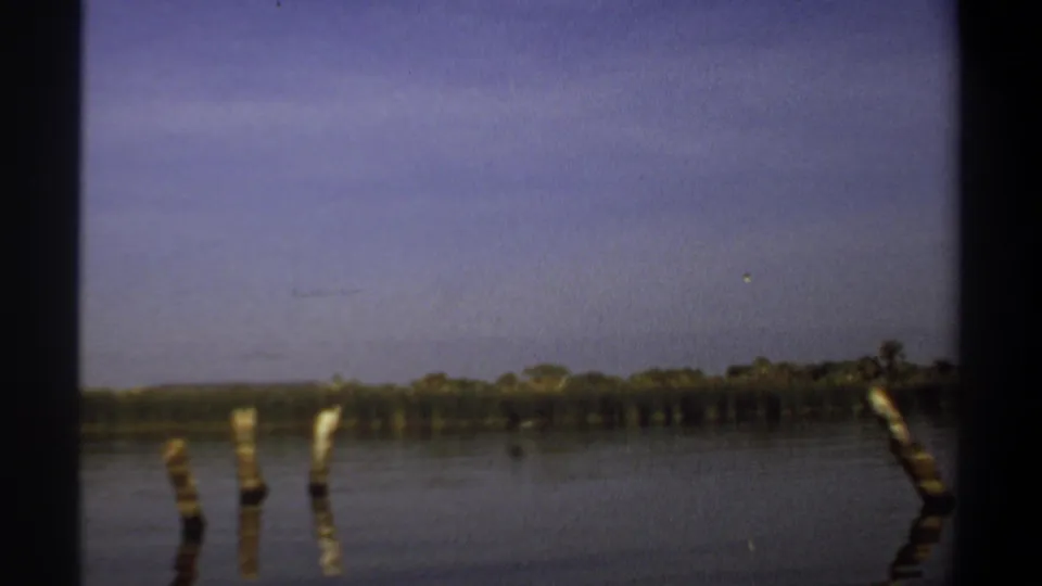 1969 Khartoum-area home-movie frame showing calm river water, wooden posts, a distant treeline, and hazy blue sky.