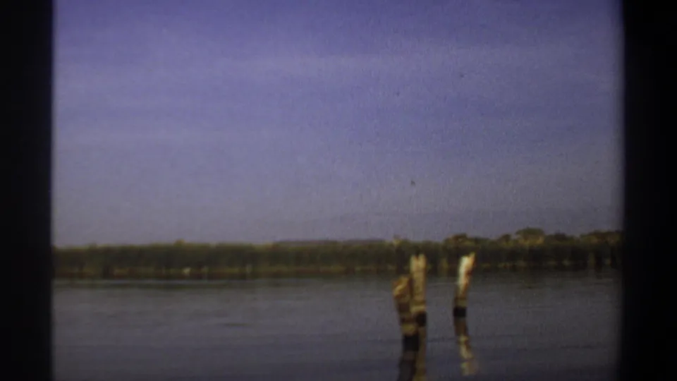 1969 Khartoum-area river view with upright wooden posts in the water, distant green vegetation, and a broad hazy sky.