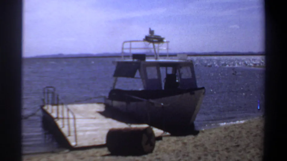 1969 Khartoum 8mm frame showing a small motorboat beside a simple wooden pier on a sandy riverbank under open sky.