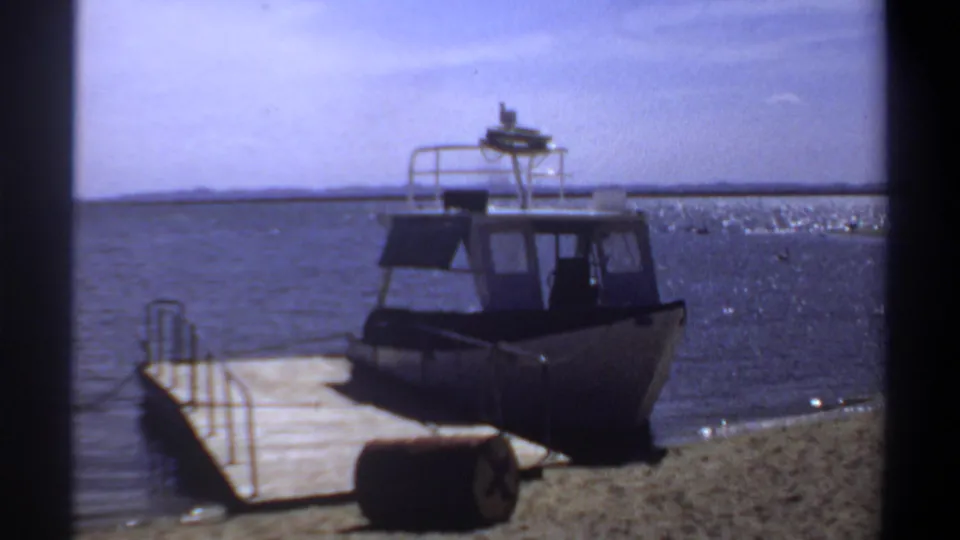 1969 Khartoum home-movie frame of a docked motorboat, simple pier, sandy bank, and broad river stretching to the horizon.