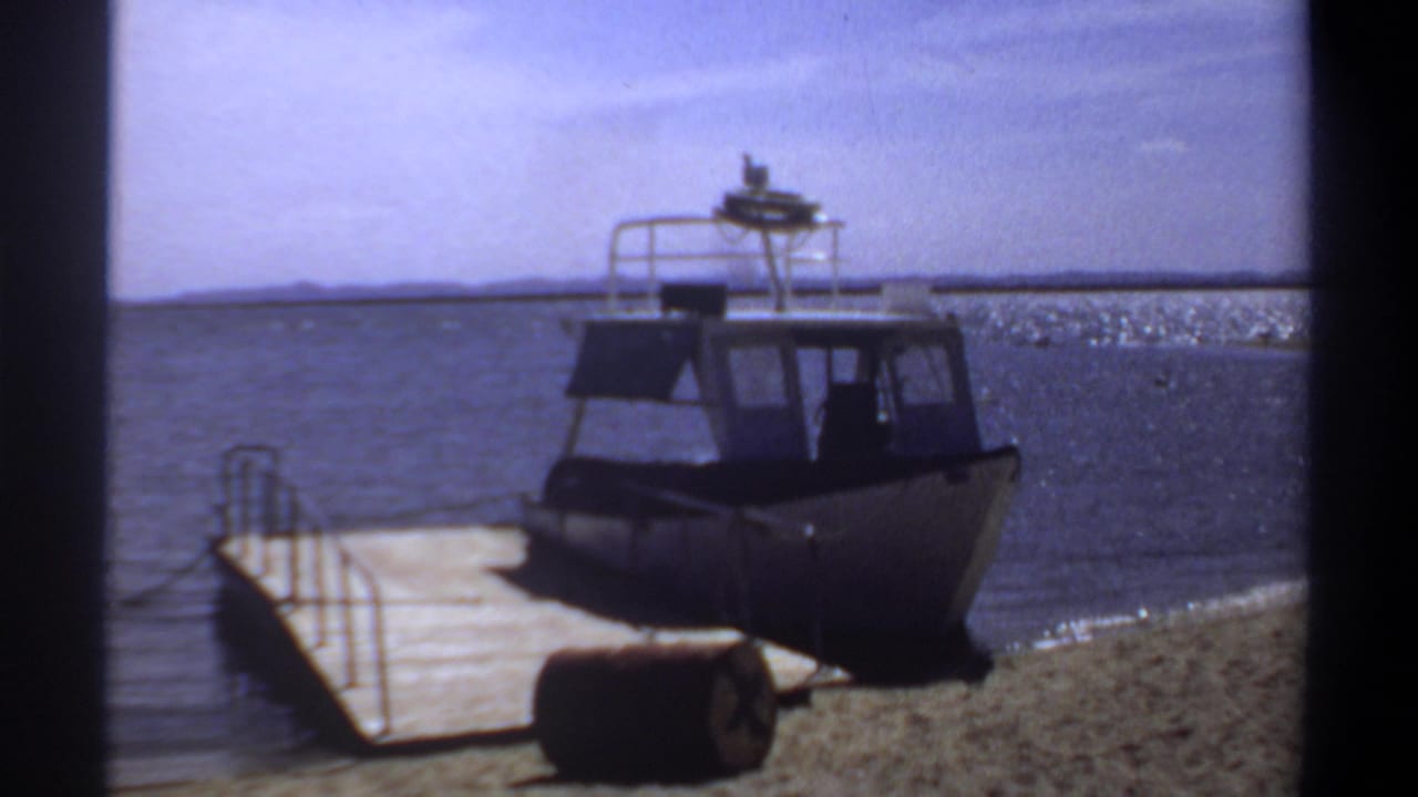 1969 Khartoum home-movie frame showing a small cabin motorboat beside a wooden pier, with a barrel on the sand and sunlit water beyond.