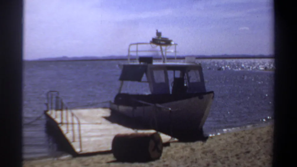 1969 Khartoum frame with a docked cabin boat, wooden pier, sandy bank, dark barrel, and sparkling water under a bright sky.
