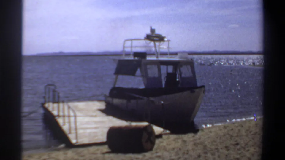 1969 Khartoum home-movie frame showing the boat from shore, with the wooden dock at left, a barrel in front, and rippling water behind.