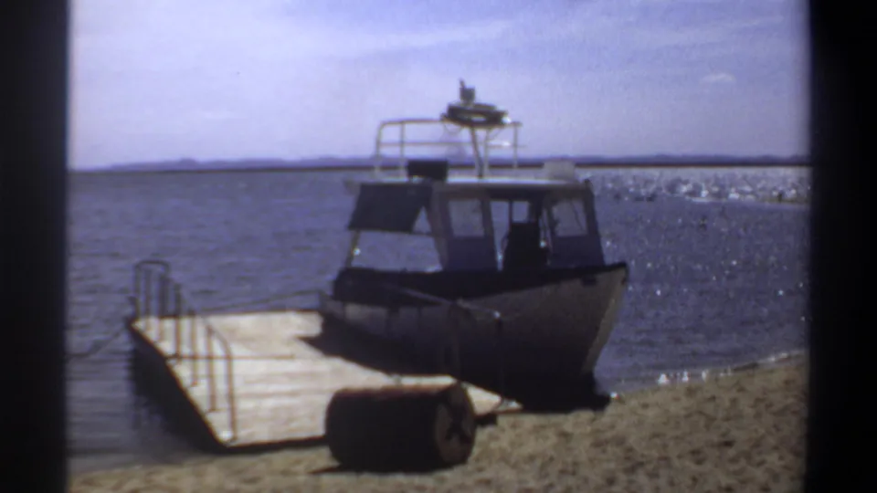 1969 Khartoum frame showing a small cabin boat moored by a wooden pier, with a barrel on shore and gentle waves along the sand.
