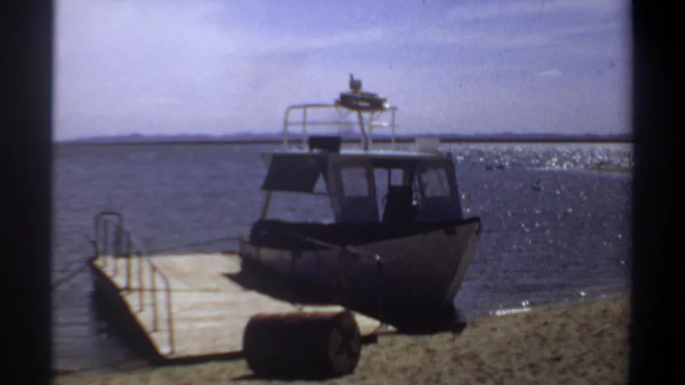 White cabin powerboat near a wooden dock on the Khartoum waterfront in 1969, with open water and a sandy bank visible.