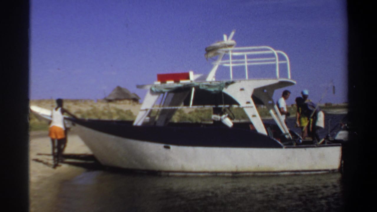 1969 Khartoum home-movie frame showing a beached white and black speedboat with several people gathered near the stern and shore.