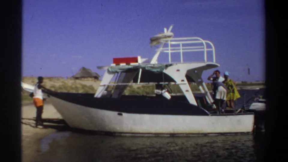 1969 Khartoum home-movie still showing a small speedboat on the shore while people stand near the rear boarding area.