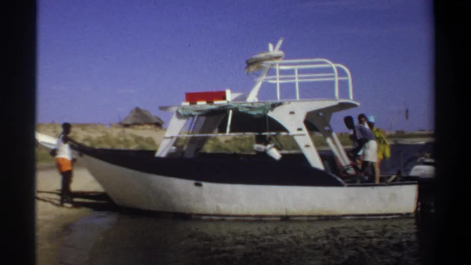 1969 Khartoum frame showing adults beside a white and black speedboat on a sandy waterside, with dark film borders visible.