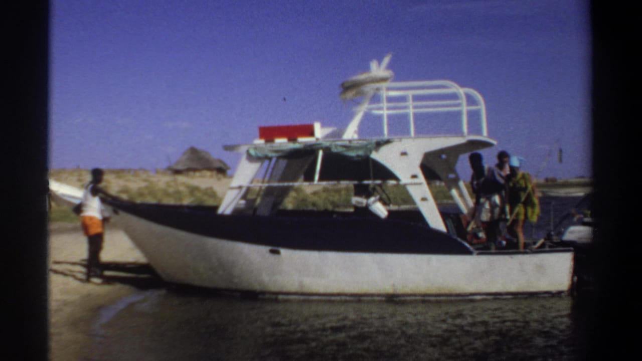 1969 Sudan home-movie frame showing a motorboat at the waterline, with people on shore and near the stern under a clear sky.
