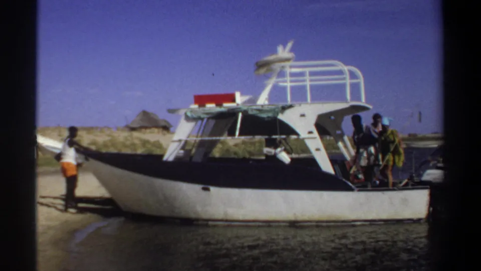 1969 Sudan home-movie frame showing a motorboat at the waterline, with people on shore and near the stern under a clear sky.