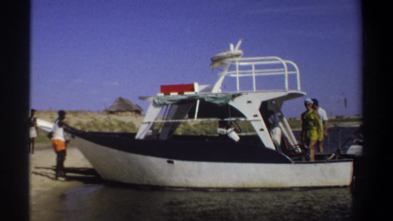 1969 Khartoum-attributed frame of a moored motorboat, with people positioned at the bow and stern on a sandy beach.