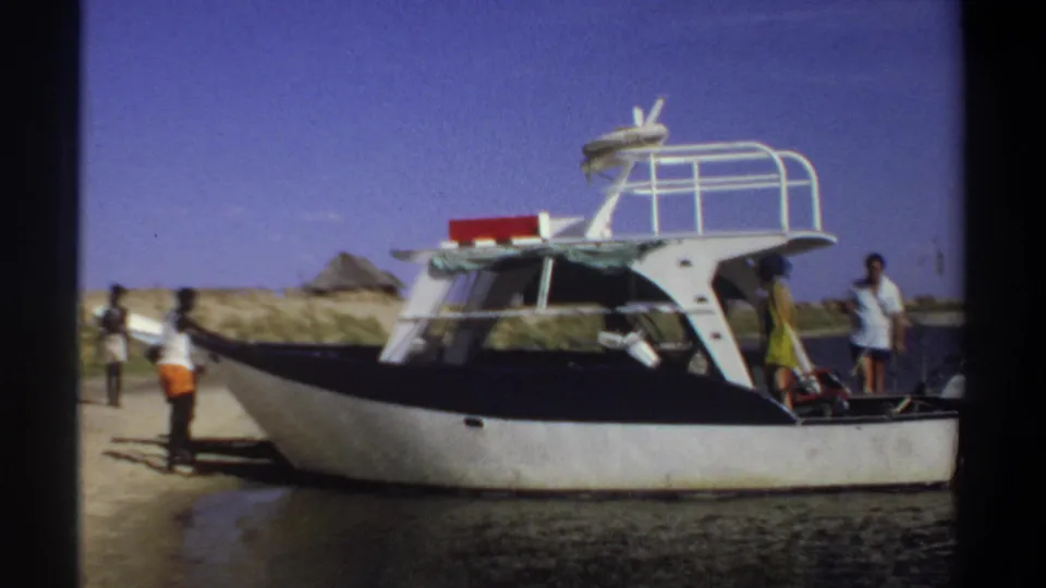 1969 Sudan travel-film frame showing people around a black-and-white motorboat on a sandy shoreline with a hut behind it.
