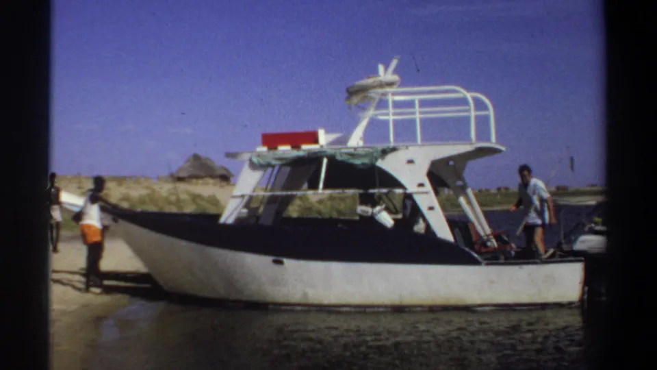 1969 Khartoum-attributed frame showing a motorboat at the shoreline, with people visible beside the vessel and dunes beyond.