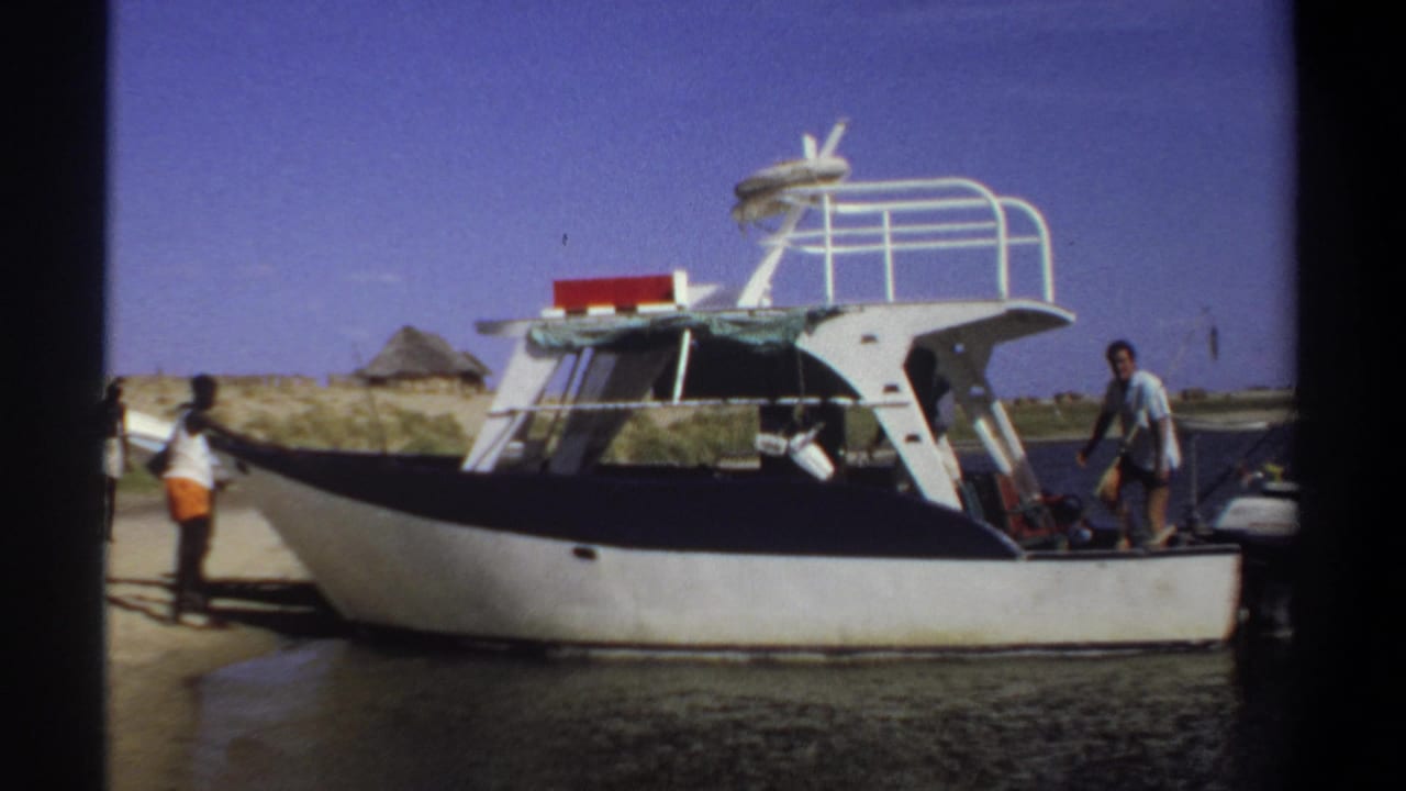 1969 Sudan home-movie frame showing a small cabin boat at shore, with people near the bow and a man standing on the deck.