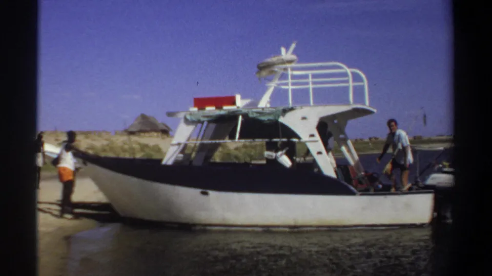 1969 Sudan home-movie frame showing a small cabin boat at shore, with people near the bow and a man standing on the deck.