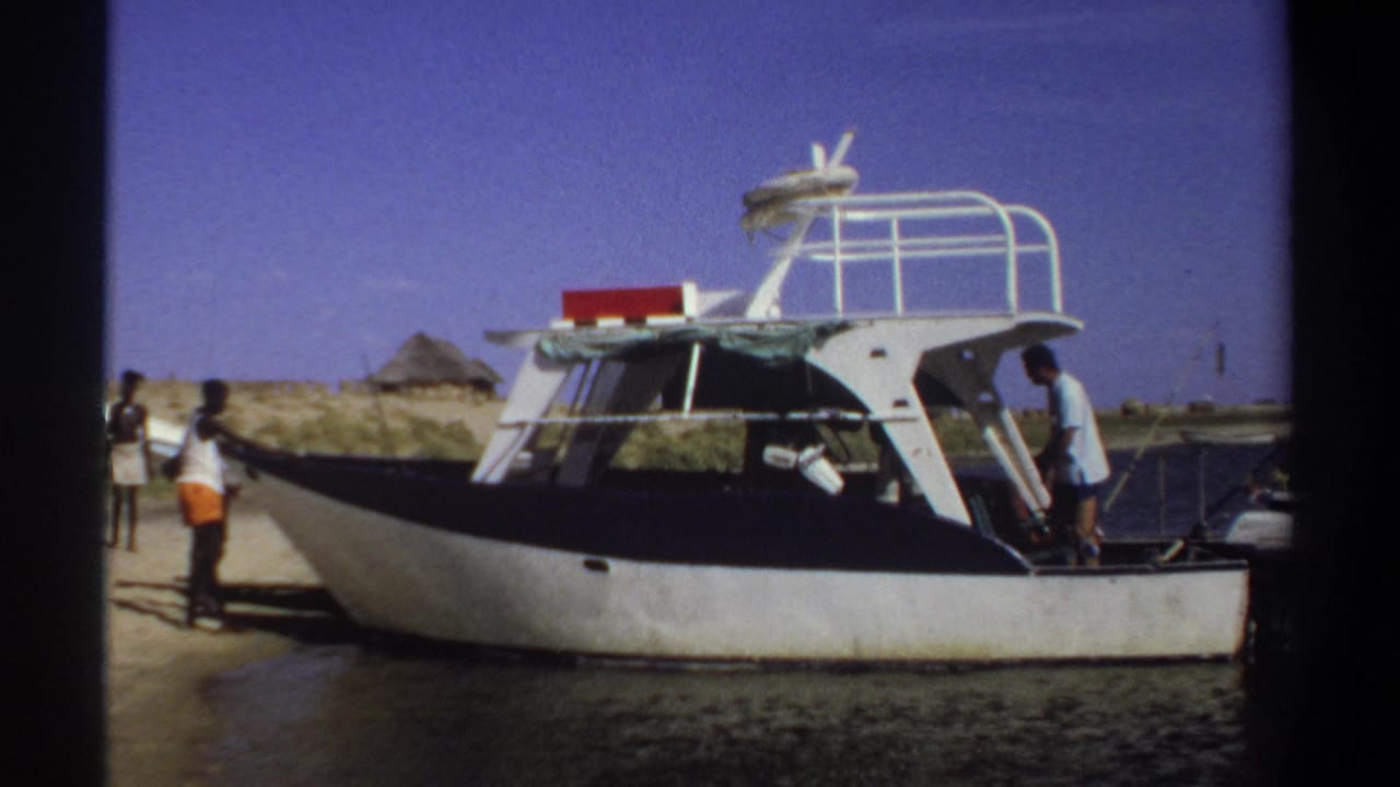 Cabin boat at shore with two people nearby and a low hut in the background.