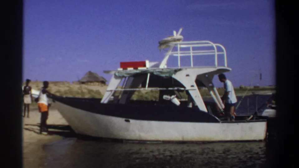 1969 Sudan shoreline view of a small cabin boat, people standing near it, and a thatched hut beyond the sand.