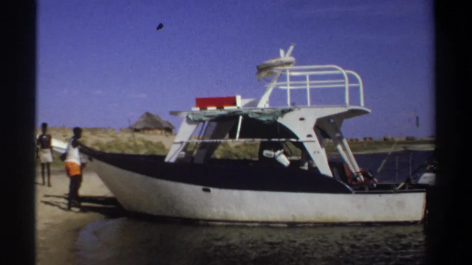 1969 Sudan home-movie frame of a cabin boat along a sandy bank, with shoreline figures and a thatched hut in the background.