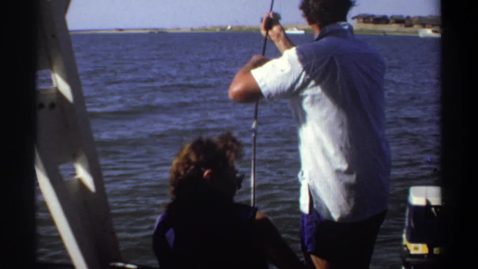 1969 Sudan home-movie frame showing a man holding a fishing line from a boat, with a seated passenger beside him on open water.