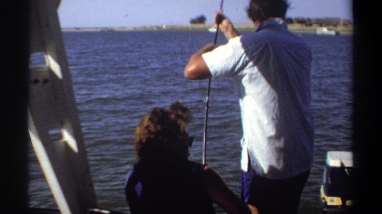 Man standing on a boat reeling a fishing rod while a seated companion faces the water.