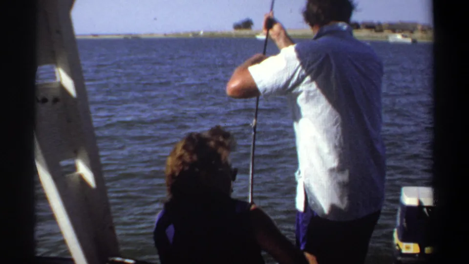 1969 Khartoum home-movie frame of a man fishing from a boat with a seated companion, ladder, water, and low shoreline visible.