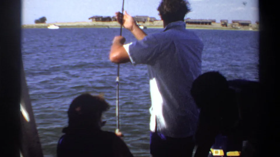 Final 1969 Khartoum boating frame with the angler reeling near a cooler, open water, shoreline structures, and a shadowed companion.