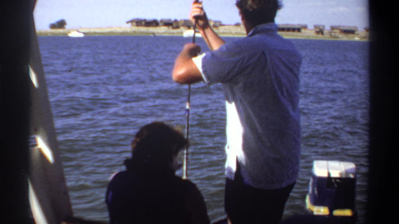 Man fishing from a boat while another person sits in shadow beside rippled lake water.