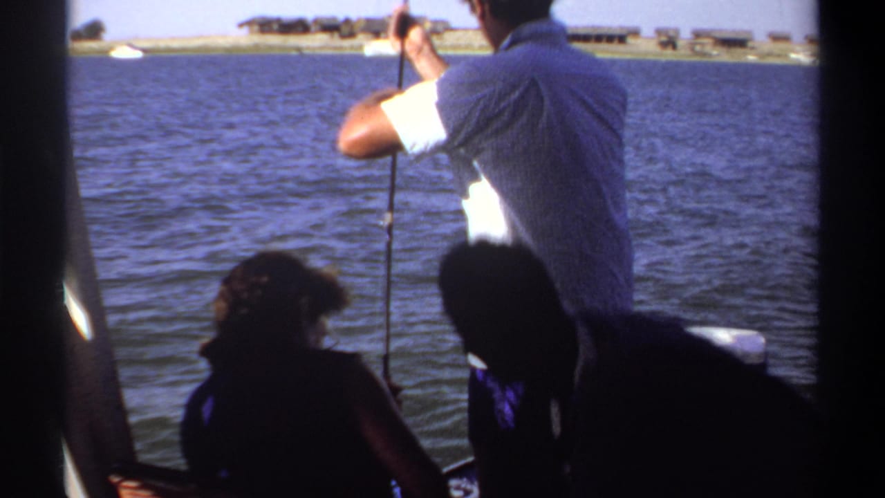 1969 Khartoum lake scene with a standing fisherman, seated companions, boat structure, and distant low shoreline buildings.