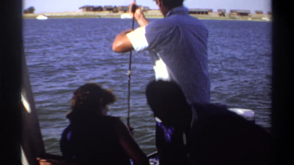 1969 Khartoum lake scene with a standing fisherman, seated companions, boat structure, and distant low shoreline buildings.