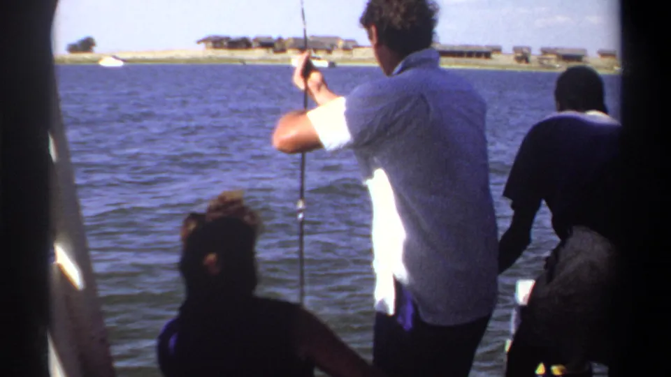 1969 Khartoum 8mm frame of a man fishing from a boat, with a foreground passenger, cooler, lake water, and shoreline beyond.