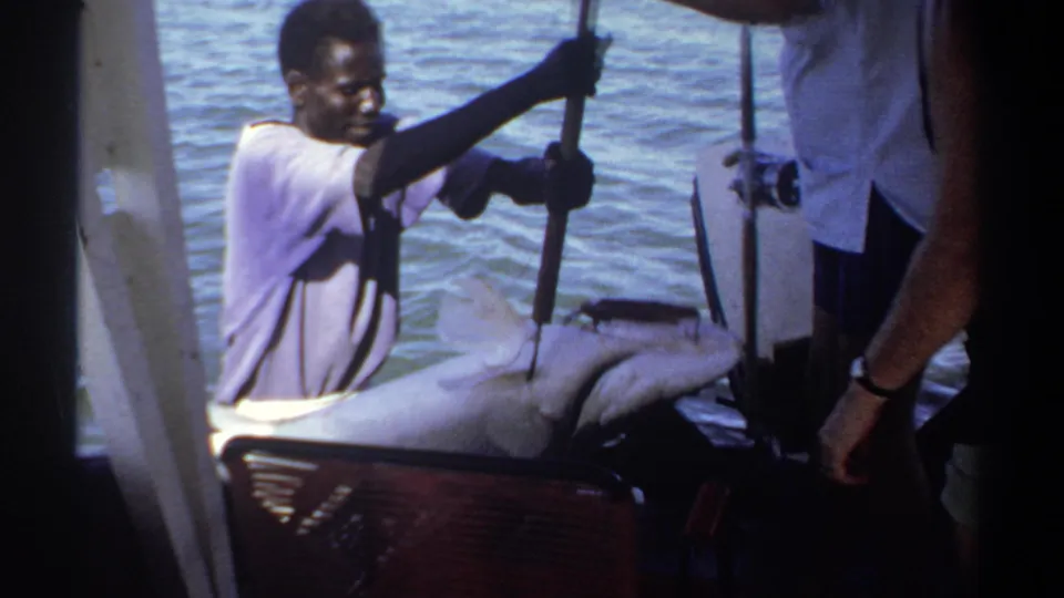 1969 Khartoum home-movie frame showing a man using a pole near a large fish on a small boat deck, with water behind him.