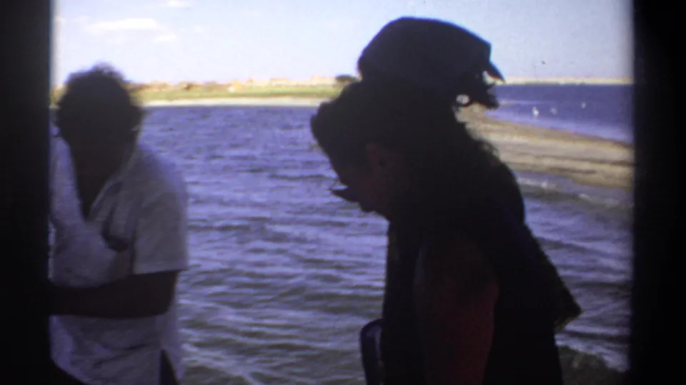 1969 Sudan 8mm frame showing two adults aboard a boat, one in a light shirt and one in dark clothing, with shoreline beyond.