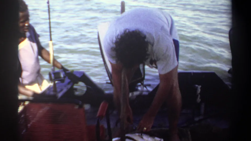 1969 Khartoum fishing footage shows an adult bending over a large fish as another person sits nearby holding a rod.