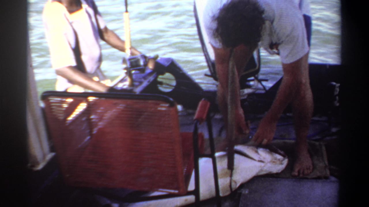 1969 Khartoum home-movie frame showing men on a small fishing boat with a large fish on deck, rod, chair, and water nearby.