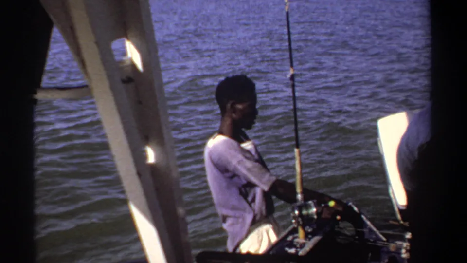 1969 Khartoum home-movie frame showing a man by the water with a fishing rod on a small boat in bright daylight.