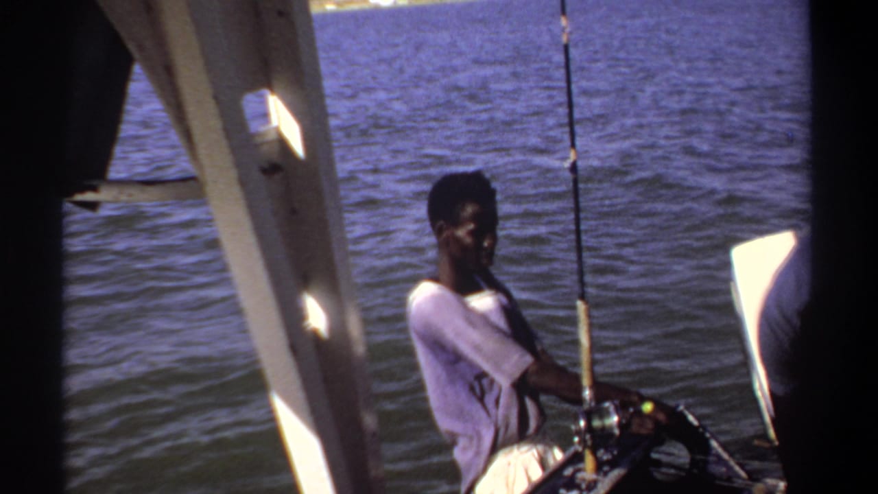 Man seated in a small boat holding a fishing rod beside calm water.