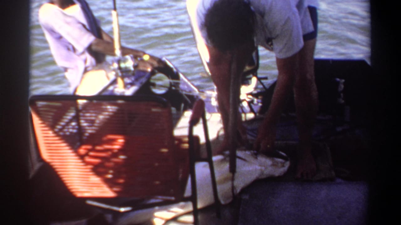 Men on a boat near fishing rods, with a red woven chair in the foreground.