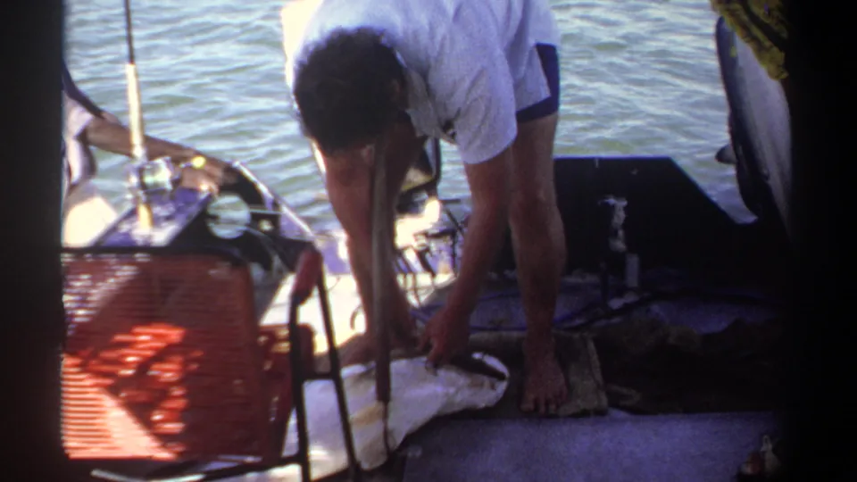 1969 Khartoum home-movie frame showing a man bending over a fish laid on a white cloth aboard a boat.