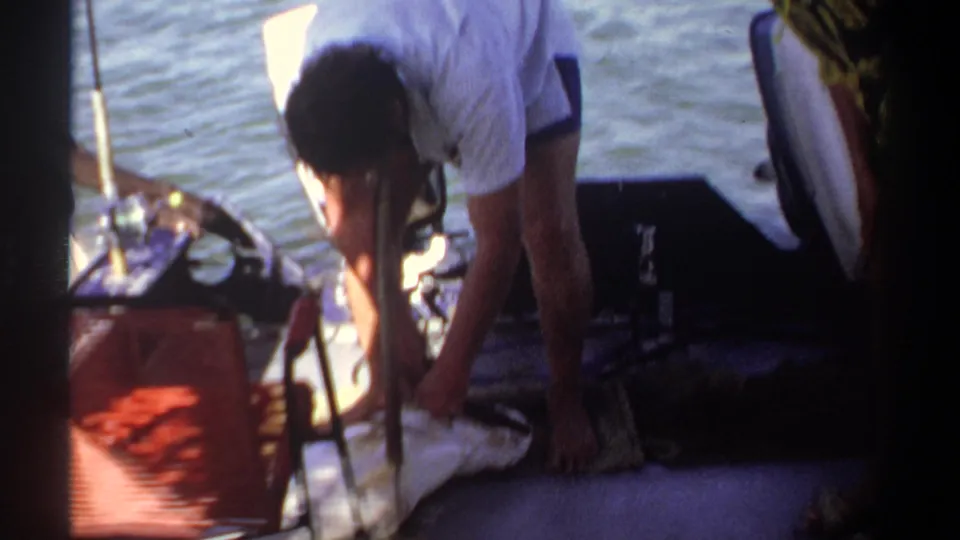 1969 Khartoum home-movie frame with a man handling a fish or gear on a boat deck, water visible behind him.