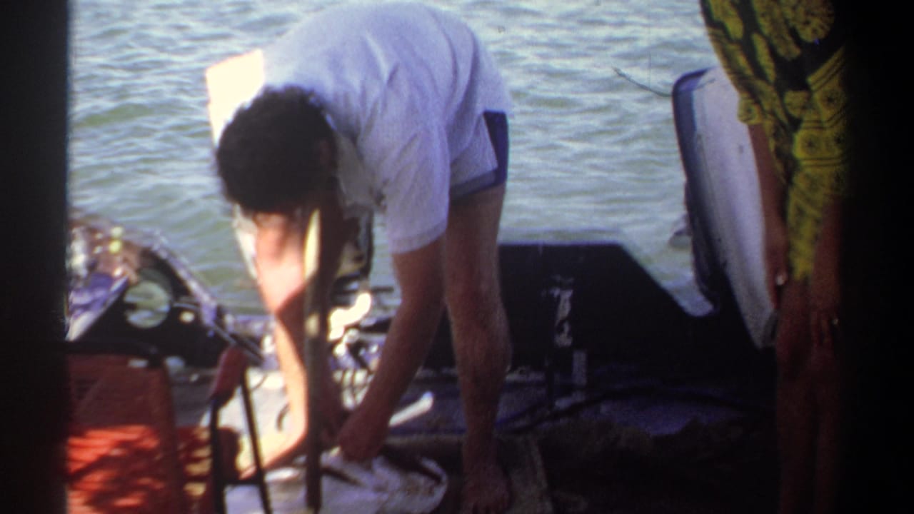 Man in a white shirt bends over gear on a small boat beside river water.