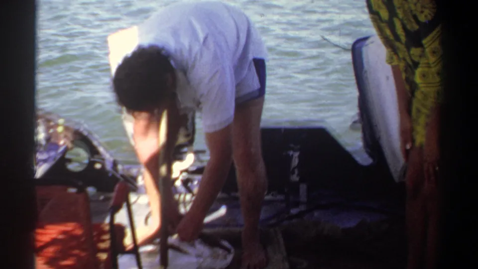 In 1969 Khartoum footage, a man in a white shirt and shorts bends over gear on a small boat on the Nile.