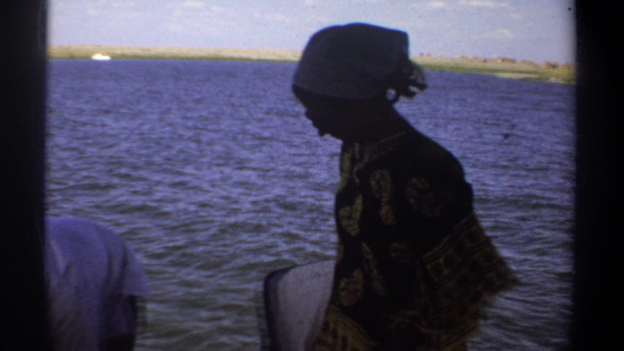 A woman in patterned clothing and a headscarf stands aboard a small boat on the Nile near Khartoum in 1969 footage.