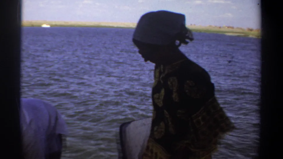 A woman in patterned clothing and a headscarf stands aboard a small boat on the Nile near Khartoum in 1969 footage.