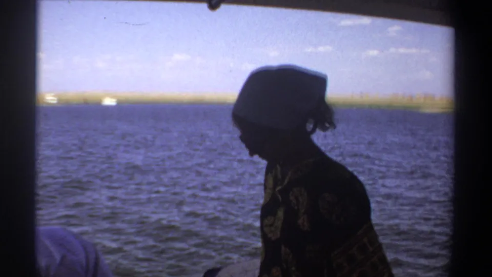 In the Khartoum boat scene, a woman in a headscarf appears in profile against rippling Nile water and a low shoreline.